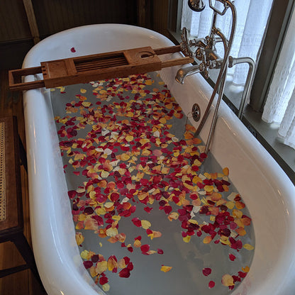 Bulk Sunset Serenade warm-toned rose petals for outdoor weddings – Prime Petal Company. Bath tub filled with water and colorful flower petals, with a wooden bath tray.