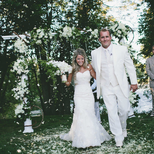 Bulk ivory rose petals for elegant wedding aisle and event décor – Prime Petal Company. Wedding couple walking down an aisle with floral decorations and guests in the background.