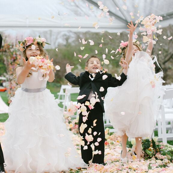 Bulk ivory cream blush rose petals for soft-toned wedding décor – Prime Petal Company. Children in formal attire are throwing flower petals at a wedding ceremony.