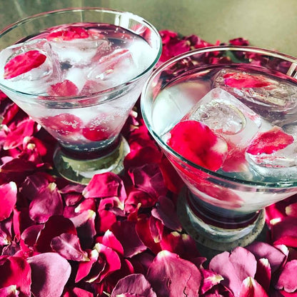 Bulk romance red rose petals for romantic wedding décor – Prime Petal Company. Two glasses with pink rose petals and ice cubes on a bed of red rose petals.