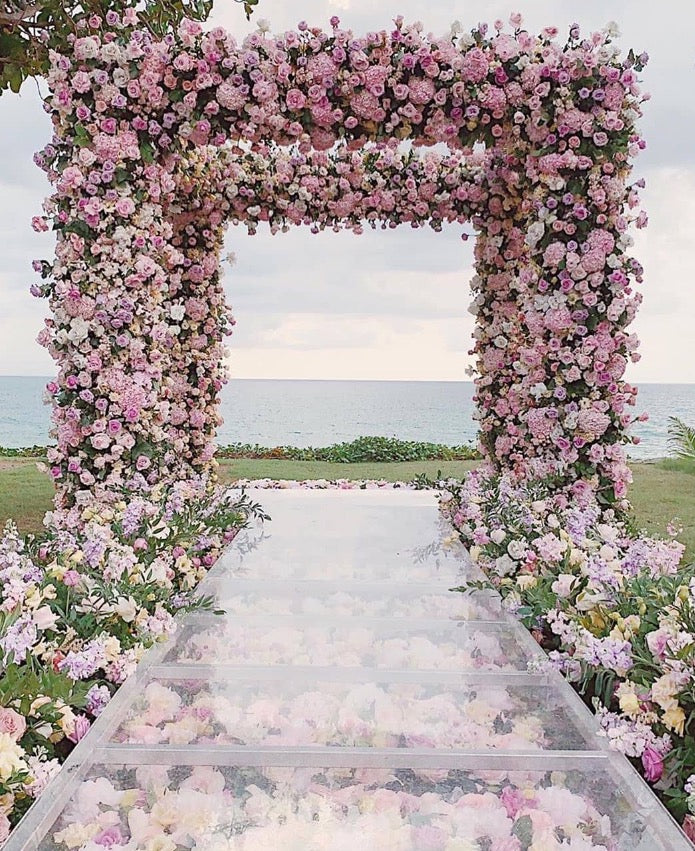 Bulk pink rose petals for wedding aisle and décor – Prime Petal Company. Floral archway with pink and white flowers leading to a clear pathway with flower petals on a grassy field.