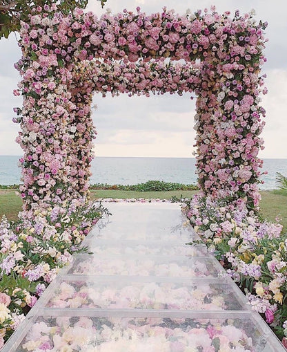 Bulk pink rose petals for wedding aisle and décor – Prime Petal Company. Floral archway with pink and white flowers leading to a clear pathway with flower petals on a grassy field.