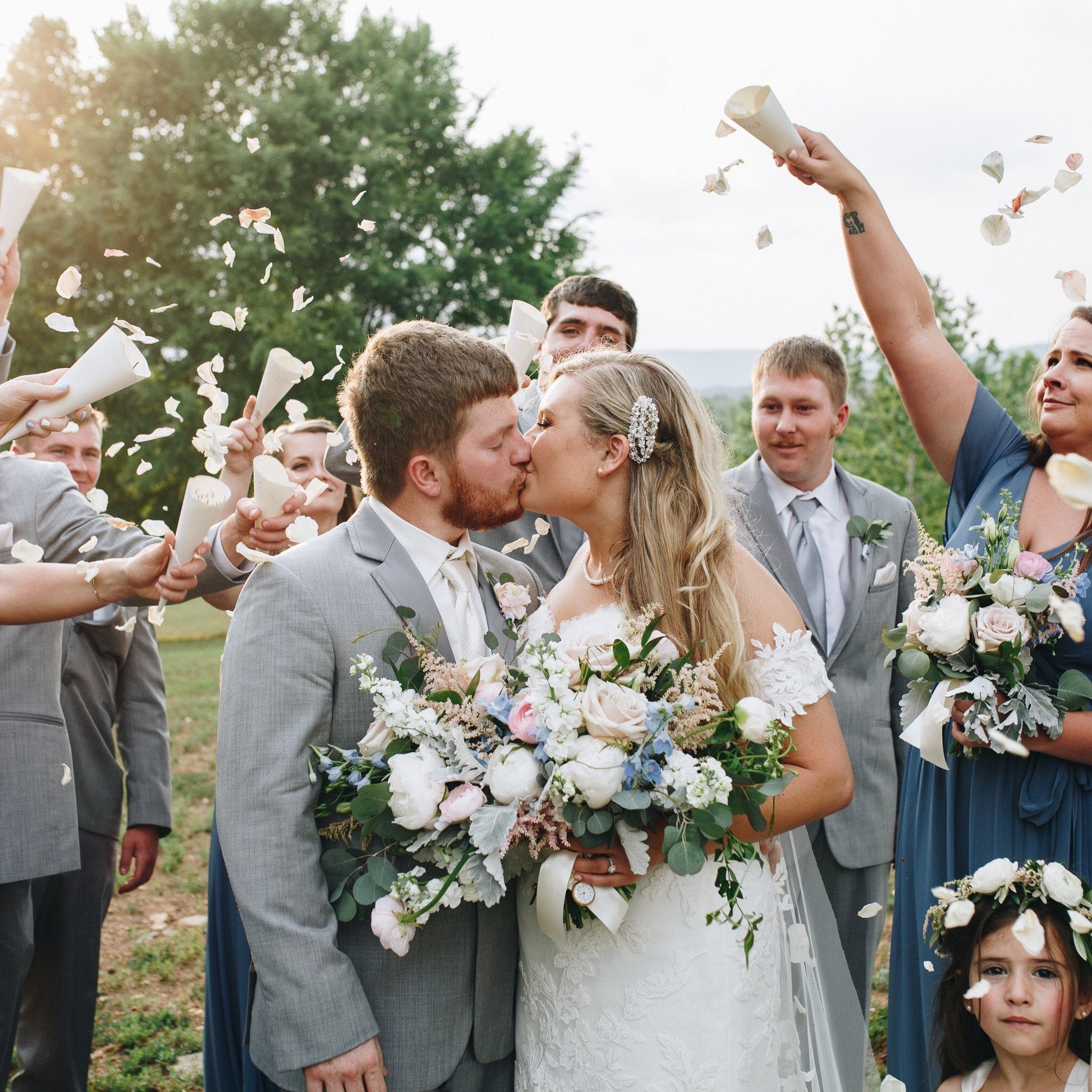 Bulk ivory cream blush rose petals for soft-toned wedding décor – Prime Petal Company.  Wedding couple kissing with confetti and guests holding bouquets outdoors.