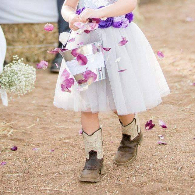 Bulk pink rose petals for wedding aisle and décor – Prime Petal Company.  Flower girl in a white dress tossing purple rose petals and cowgirl boots standing on the outdoor wedding pathway.