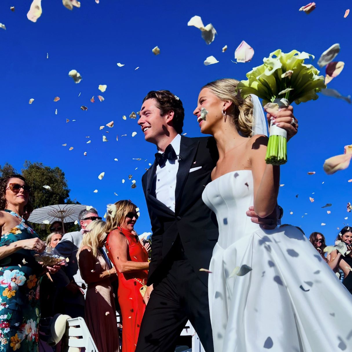 Bulk pink rose petals for wedding aisle and décor – Prime Petal Company. A Wedding couple standing under rose petals confetti against a blue sky with guests in the background.
