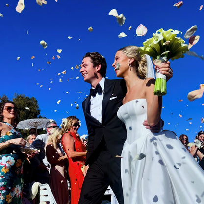 Bulk pink rose petals for wedding aisle and décor – Prime Petal Company. A Wedding couple standing under rose petals confetti against a blue sky with guests in the background.