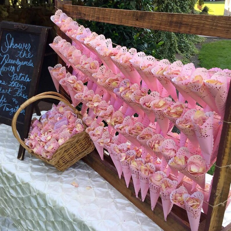 Bulk Blissful Bloom mixed flower petals for weddings and events – Prime Petal Company. Pink decorative cones filled with pink rose petals with lace details on a wooden stand outdoors.