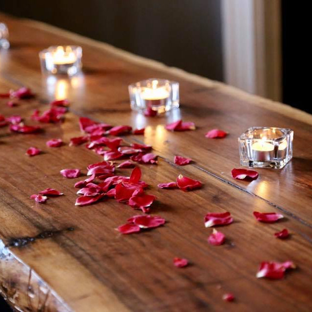 Bulk ruby rose petals for weddings and romantic events – Prime Petal Company. rose petals scattered on a wooden bar with small candles.