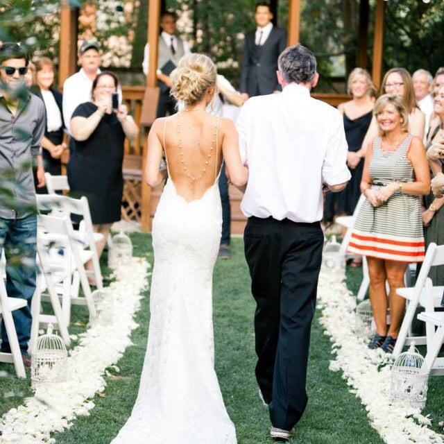 Bulk ivory cream blush rose petals for soft-toned wedding décor – Prime Petal Company. Wedding ceremony with a bride walking down the aisle lined with ivory rose petals, with her father, surrounded by guests under a wooden gazebo.