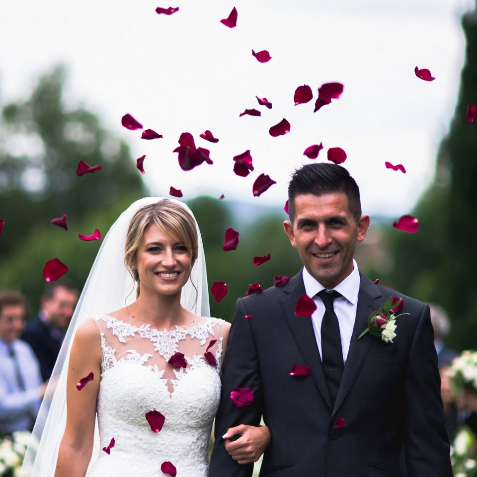 Bulk crimson rose petals for rich red wedding and event décor – Prime Petal Company. Wedding couple standing outdoors with red rose petals falling around them.